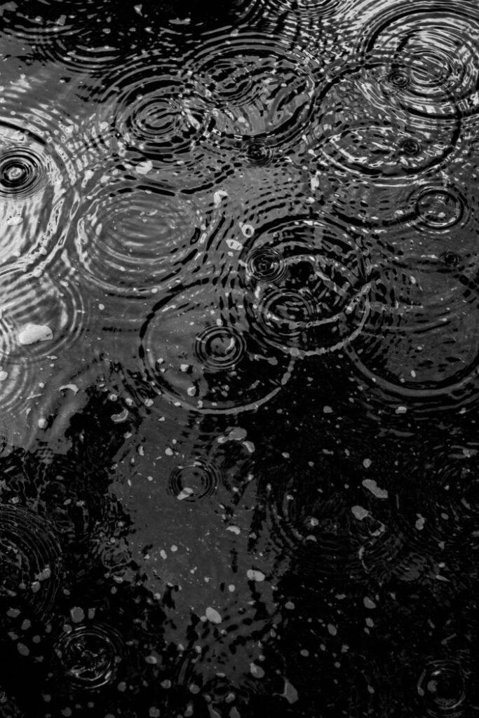 Monochrome image of raindrops creating ripples on a water surface.